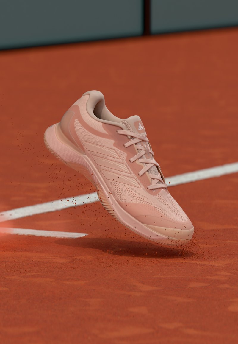 Hellrosa Sportschuh mit Mesh-Obermaterial, strukturierten Überzügen und Schnürdesign, schwebt über einem Sandplatz mit verstreutem Staub.