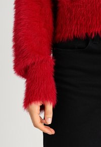 Czerwony, rozciągliwy, krótki sweter z długimi rękawami, o teksturowanej, puszystej powierzchni. Noszony na czarnej, wysokiej spódnicy.