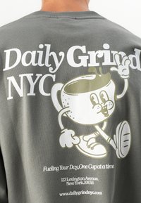 Gray sweatshirt with "Daily Grind NYC," a smiling coffee cup character waving, and address and website text below.