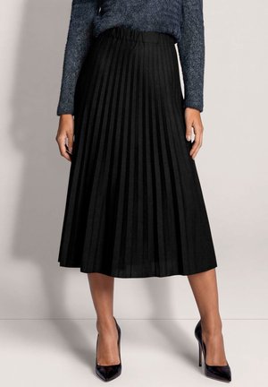 Femme portant une jupe midi plissée noire, un pull pelucheux bleu foncé et des chaussures à talons noires, debout devant un fond uni.