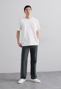 T-shirt branca de algodão, de corte confortável; calças cinzas escuras com riscas laterais brancas; usadas com ténis brancos; de pé contra um fundo neutro.