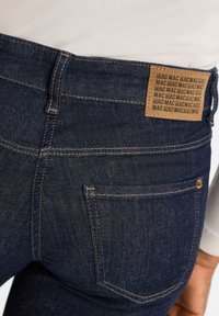 Jean en denim bleu foncé avec une finition texturée, présentant une étiquette en cuir marron embossée avec "MAC" et des coutures contrastantes sur les poches.