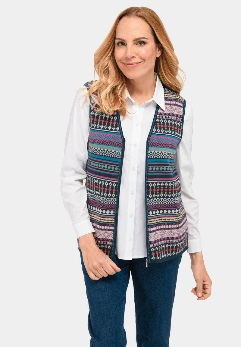Patroonvest met bloemen- en geometrische ontwerpen in blauw, roze en wit. Voorzien van een rits aan de voorkant, gedragen over een wit shirt.