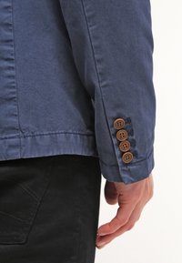 Detail námořnického modrého saka s manžetou na knoflíky, která má čtyři hnědé knoflíky, vyrobeného z hladké látky s jemnou texturou a prošíváním.