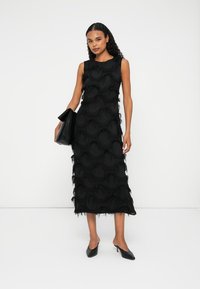 Mujer de pie en un vestido midi negro sin mangas con patrones circulares texturizados, sosteniendo un bolso negro de mano y usando mules negros con tacón.