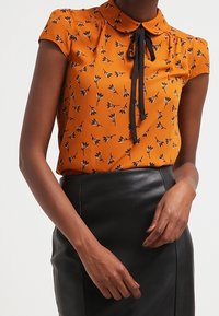 Blusa arancione a maniche corte con motivo floreale nero, colletto e dettaglio con fiocco, abbinata a una gonna nera in pelle a vita alta.
