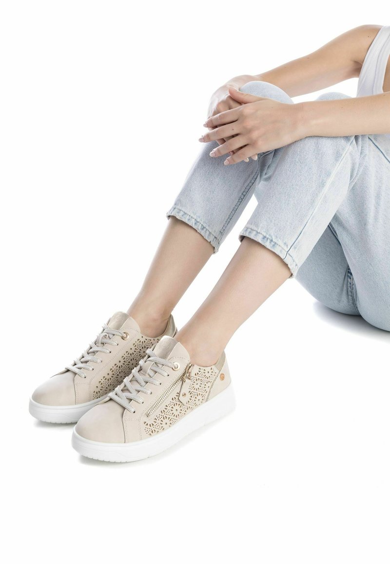 Zapatillas beige con diseño perforado, que cuentan con una cremallera lateral, tela texturizada, suela blanca y punta redonda. Combinadas con denim claro.