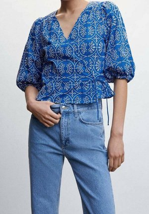 Femme portant un chemisier bleu à motifs avec col en V et manches bouffantes, ainsi qu'un jean taille haute bleu clair, main reposant près de la poche.