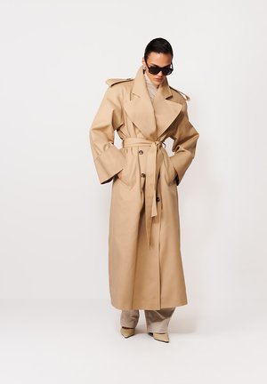 Žena v dlhom béžovom trenčkotom kabáte so širokými klopami, zoštíhleným pásom, nadrozmernými rukávmi, slnečnými okuliarmi a svetlými špicatými topánkami.