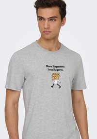 Γκρι βαμβακερό μπλουζάκι με κοντά μανίκια που απεικονίζει ένα σχέδιο με χαρακτήρα που μεταφέρει μπαγκέτες και το κείμενο «Περισσότερες Μπαγκέτες, Λιγότερες Μετανοιώσεις.»