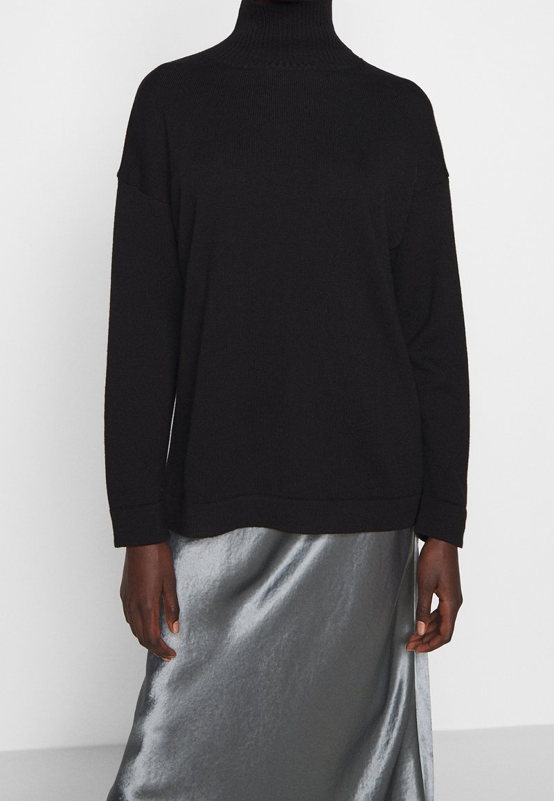 Pull col roulé noir à manches longues porté avec une jupe en satin argenté brillant, présenté du cou jusqu'à mi-cuisse sur un fond uni.