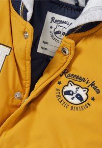 Żółta ocieplana kurtka z granatowym podbiciem, metalowymi zatrzaskami i haftowanym logo szopa. Posiada szary kołnierz wyłożony polarem.