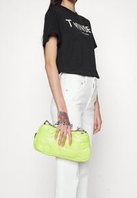 Sac à main matelassé vert néon avec une poignée en chaîne argentée, tenu d'une main. Le modèle porte une chemise noire et un jean blanc avec un tatouage floral.
