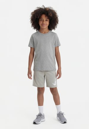 Niño con cabello rizado que lleva una camiseta gris, pantalones cortos gris claro con el logo blanco de Nike, calcetines blancos y zapatillas Nike grises, de pie y sonriendo.