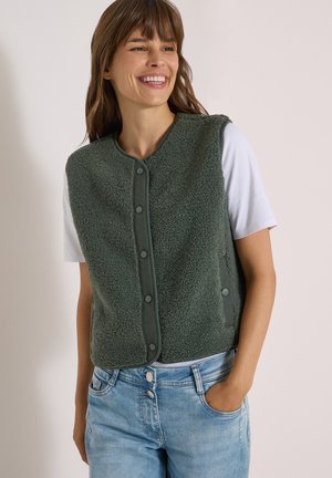Smilende kvinde iført en grøn knap-fleece vest over en hvid kortærmet skjorte og lyseblå jeans, med den ene hånd i lommen.