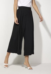 Woman wearing high-waisted black wide-leg cropped pants with pleats and white heeled open-toe mules against a plain white background.