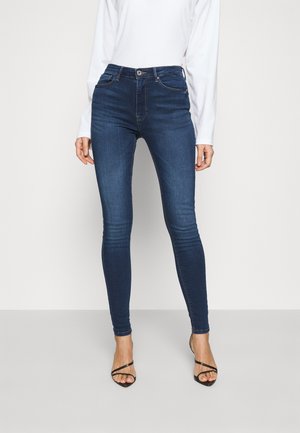 Femme portant un jean skinny bleu foncé, un top blanc à manches longues et des sandales noires à talons hauts à brides, debout devant un fond uni.