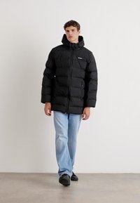 Zwarte pufferjas met een capuchon, voorzien van horizontale quiltsecties, ritszakken en een klein logo aan de linkerzijde. Gekleed met lichtblauwe jeans.