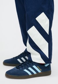 Μπλε σουέντ αθλητικά παπούτσια με τρεις ανοιχτό μπλε ρίγες, καφέ ελαστικέ�ς σόλες, σε συνδυασμό με σκούρο μπλε παντελόνια που έχουν λευκές διαγώνιες ρίγες.