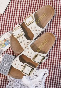 Witte sandalen met dubbele gespen en gouden studs op een rood geruit stuk stof, omringd door speelkaarten en een witte netten tas.