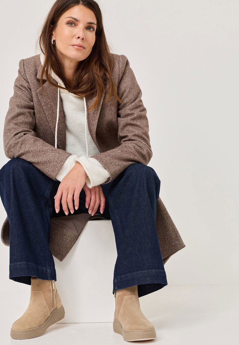 Beige ankelstøvler, herringbone brun frakke, grå hoodie og mørk denim bukser med bred ben. Glatte teksturer med kontrasterende farver.