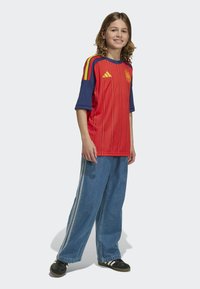 Röd och blå Adidas-tröja med korta ärmar, vertikala ränder och logotyp; kombinerad med blå vida jeans och svarta sneakers.