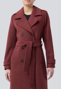 Kvinna i en dubbelknäppt vinröd ullrock med breda slag, svarta knappar, sidofickor, manschetter med remmar och ett knytbart midjebälte.