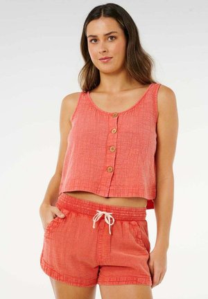 Mujer con top sin mangas texturizado de coral con botones y pantalones cortos a juego con cordón, de pie con una mano en el bolsillo contra un fondo liso.