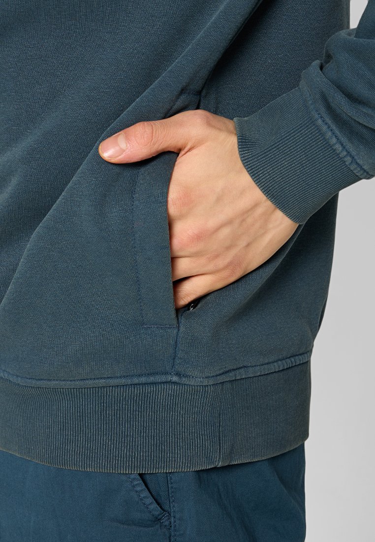 Ciemnozielony sweter z kieszenią z przodu, żebrowany przy rękawach i u dołu. Materiał wydaje się miękki, o gładkiej teksturze i minimalnych detalach szwów.