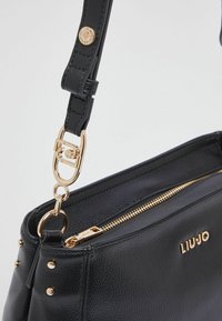 Sac à main en cuir noir avec ferrures dorées, finition texturée, longue bandoulière réglable avec boucle, et détail logo sur le devant. Fermeture à glissière.