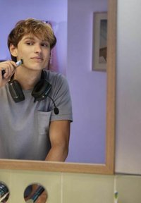 Elektrische scheerapparaat in de hand van een persoon die zwarte koptelefoon met microfoon draagt. Grijze t-shirt, badkameromgeving met een spiegel en pastelkleuren.