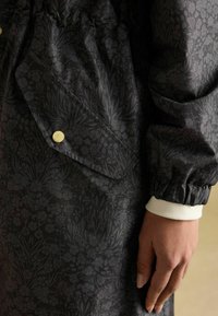 Čierna bunda s kvetinovým vzorom, obsahujúca veľkú prednú kapsu, elastické manžety a hladký povrch. Viditeľný detail so zlatým zapínaním.