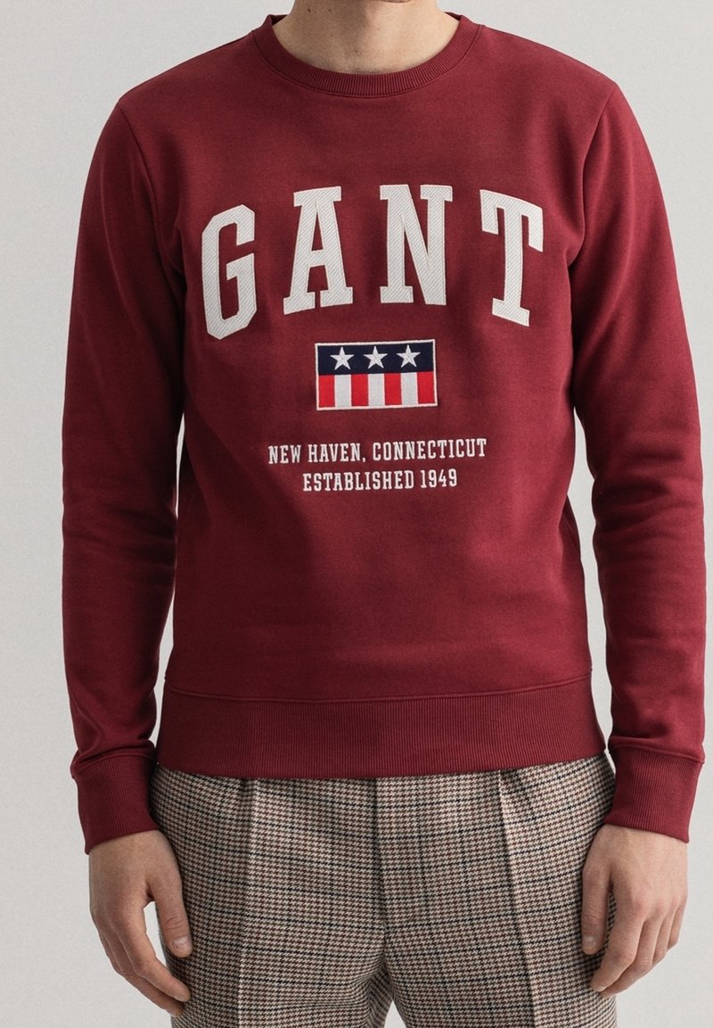 Sweatshirt bordeaux avec logo GANT, emblème étoiles et rayures, et texte "New Haven, Connecticut Established 1949,", porté avec un pantalon à carreaux.