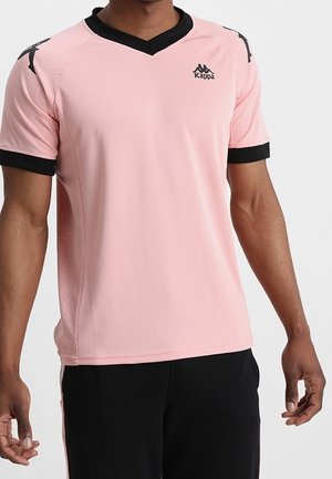 Mężczyzna ubrany w różową sportową koszulkę Kappa z czarnym kołnierzem i krawędziami rękawów, w połączeniu z czarnymi spodniami.