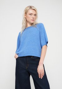 Μπλε πουλόβερ με κοντά μανίκια από απαλό πλεκτό ύφασμα, σε χαλαρή γραμμή, με στρογγυλεμένο λαιμό. Συνδυάζεται με σκούρα μπλε παντελόνια τζιν με φαρδιά μπατζάκια και πορτοκαλί ραφές.