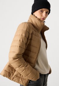 Tan pufferjakke med vandret syninger, høj krave og lynlås lukning. Båret over en beige top og parret med mørke jeans. Sort huedragt som accent.