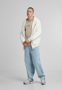 Witte zip-up hoodie, beige aansluitend tanktop, lichtblauwe losse jeans en zilveren sportschoenen met blauwe accenten. Neutrale achtergrond.
