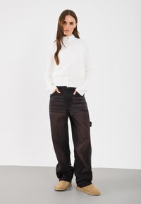 Fehér, cipzáras pulóver bordázott textúrával, fekete, bő nadrággal és bézs slip-on cipővel. A modell a zsebében lévő kezével áll egy szürke padlón.