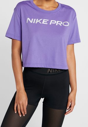 Persoon die een paarse cropped T-shirt met de tekst "NIKE PRO" draagt en zwarte shorts met hoge taille met semi-transparante zwarte legging eronder.