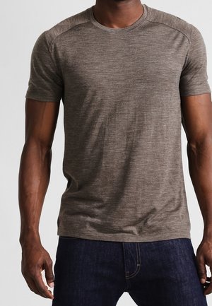 Mężczyzna ubrany w dopasowaną brązową melanżową koszulkę z krótkim rękawem i ciemnoniebieskie dżinsy, stojący z rękami swobodnie opuszczonymi wzdłuż ciała.