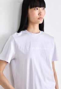 Světle fialové bavlněné tričko s krátkým rukávem a kulatým výstřihem, s logem "Calvin Klein Jeans" v bílém písmu. Hladká textura.