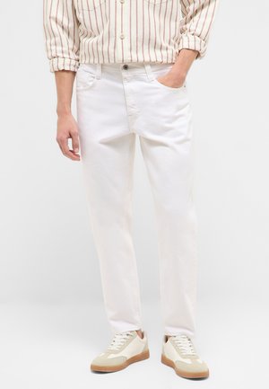 Mann trägt weiße Jeans mit einem beige gestreiften Hemd, in die Hose gesteckt, eine Hand in der Tasche und beige Sneakers mit Gummisohlen vor einfarbigem Hintergrund.