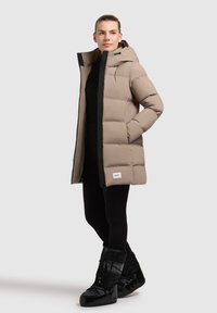 Beige színű puffertakaró kapucnival, fekete cipzárral és rugalmas mandzsettaakkal. Fekete leggings-szel viselve, fényes fekete csizmával párosítva.