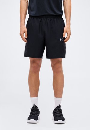 Άνδρας που φοράει μαύρα αθλητικά σορτς Under Armour, μαύρα αθλητικά παπούτσια και λευκές κάλτσες, στέκεται μπροστά σε ένα απλό φωτεινό φόντο.