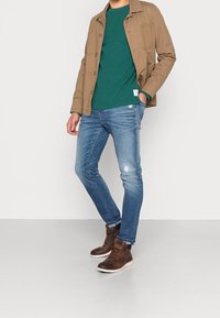 Hnědá bunda přes zelenou košili, doplněná světle modrými džíny a hnědými kotníkovými botami. Ošuntělé džíny mají jemné trhliny, což zdůrazňuje neformální design.