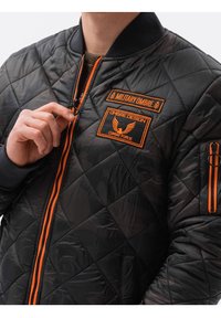 Sort quiltet bomberjakke med orange lynlås og mærker. Har ribbede ærmekanter og krave, med detaljeret syning og flere lommer.