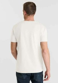 Uomo che indossa una semplice t-shirt bianca a maniche corte e jeans scuri, voltato di spalle contro uno sfondo chiaro e neutro.