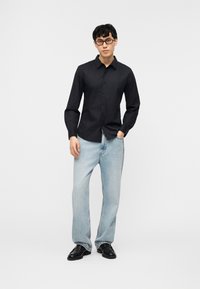 Άνδρας που φοράει μαύρο πουκάμισο με κουμπιά, ανοιχτογάλατσα τζιν, μαύρα παπούτσια και γυαλιά, στέκεται με το ένα χέρι στην τσέπη μπροστά σε απλό φόντο.