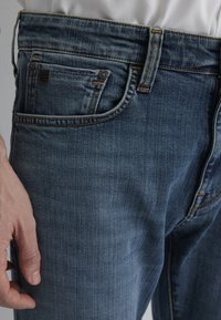 Denim jeans in medium blue with subtle fading, multiple pockets, and contrasting orange stitching. Material is textured and durable.