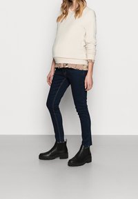Sweatshirt beige clair avec des poignets côtelés, chemise à motifs floraux en dessous, jeans skinny bleu foncé avec des accents zippés, et bottines noires.